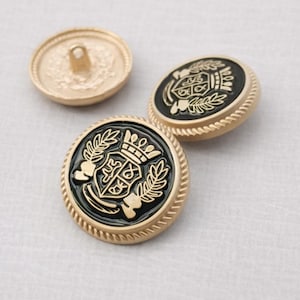 Pode incluir: Close-up de três botões dourados com um emblema preto. Os botões apresentam um design detalhado com uma coroa, um escudo e folhas de louro. Os botões são redondos com uma borda texturizada e uma presilha na parte de trás.