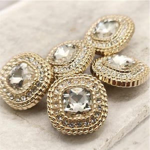 Peut inclure: Gros plan sur cinq boutons dorés avec des pierres centrales en cristal clair, de forme carrée et ovale. Chaque bouton est entouré d'un anneau de petits cristaux transparents. Les boutons sont disposés sur une surface claire.