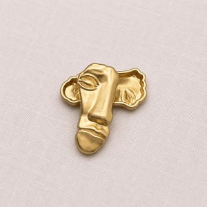 May include: A gold-toned metal brooch featuring an abstract, stylized human face design. The piece displays a smooth, polished surface with recessed areas that define the eye, nose, and mouth contours, creating a sculptural, relief-like appearance.