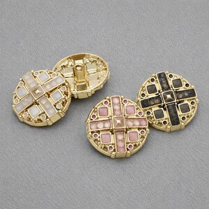 May include: Close-up of decorative gold buttons with cross-shaped designs. The buttons feature various colors, including white, pink, and black, with small, round accents. The buttons are set against a gray background.