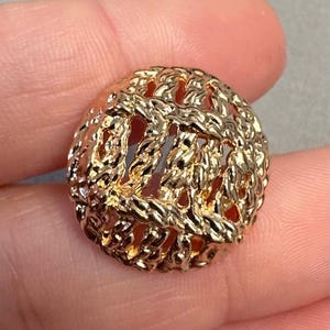 Könnte beinhalten: Ein goldfarbener, runder Knopf mit einem strukturierten, gewebten Design. Der Knopf hat eine Reihe von Öffnungen im gesamten Design. Der Knopf wird in der Hand einer Person gehalten.