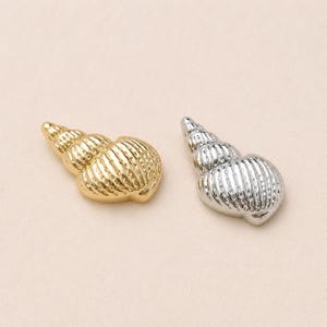 Puede incluir: Dos dijes en forma de caracola, uno con acabado en tono dorado pulido y el otro en tono plateado pulido. Cada caracola presenta una forma cónica en espiral con crestas verticales detalladas y una superficie metálica lisa y reflectante.