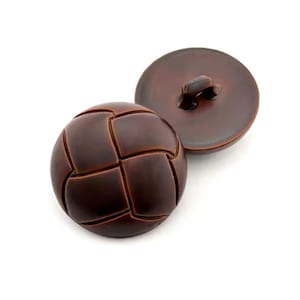 May include: Two brown buttons with a textured, stitched design. One button is round with a loop on the back, and the other is a sphere with a grid pattern. The buttons are a dark brown color.