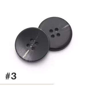 K&ouml;nnte beinhalten: Zwei schwarze, runde Kn&ouml;pfe mit jeweils vier L&ouml;chern. Die Kn&ouml;pfe haben einen leichten Marmoreffekt und sind vor einem wei&szlig;en Hintergrund platziert. Die Nummer "#3" ist unten links in Schwarz aufgedruckt.