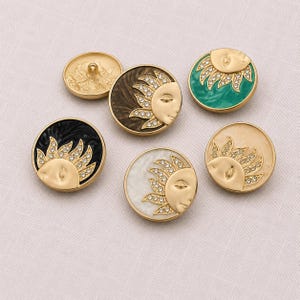 Peut inclure: Un ensemble de cinq boutons circulaires présentant un motif de visage de soleil doré avec des cristaux clairs incrustés sur les rayons. Les boutons ont des fonds en émail coloré noir, blanc, crème, marron et bleu sarcelle, un bouton montre le dos.