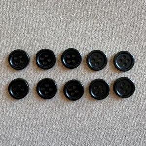 Könnte beinhalten: Zehn schwarze Plastikknöpfe mit jeweils vier Löchern. Die Knöpfe sind in zwei Reihen zu fünf angeordnet.