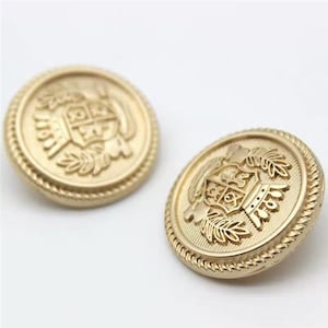 May include: Two gold-colored, round buttons with a detailed embossed design. Each button features a central crest design surrounded by laurel leaves and a rope-like border. The buttons are likely for clothing or decorative purposes.