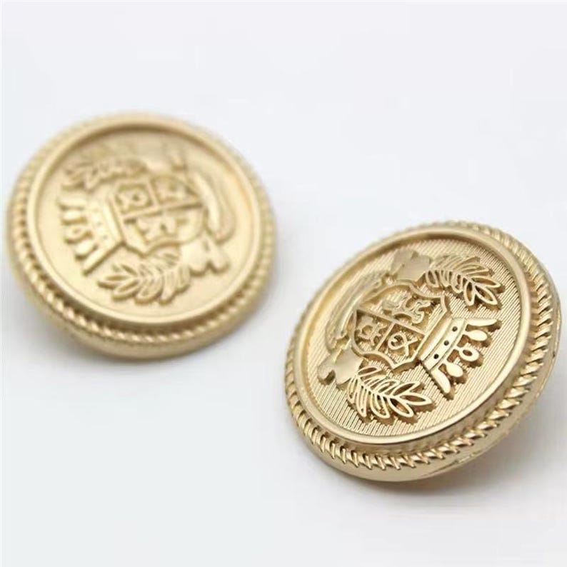 May include: Two gold-colored, round buttons with a detailed embossed design. Each button features a central crest design surrounded by laurel leaves and a rope-like border. The buttons are likely for clothing or decorative purposes.