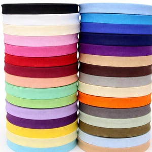 以下が含まれることがあります： 黒、白、ピンク、青など、さまざまな色合いのカラフルな布バイアステープのスタック。 テープはきちんと巻かれて積み重ねられており、縫製やクラフトプロジェクトに適したさまざまな色が表示されています。 画像は明るく照らされており、質感と色合いが強調されています。