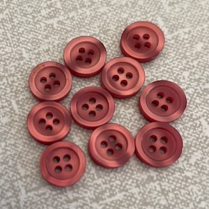 Può includere: Dieci bottoni di plastica rosa con quattro fori ciascuno. I bottoni sono disposti a semicerchio su una superficie grigio chiaro.