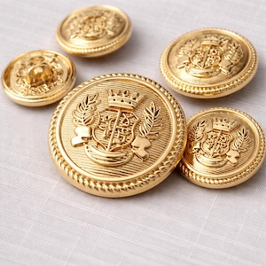 Puede incluir: Una colección de botones metálicos dorados con intrincados diseños en relieve. Los botones presentan un motivo de corona, escudo y corona de laurel. Los botones varían en tamaño y están dispuestos sobre una tela de color claro.