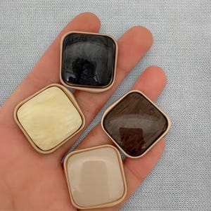 Könnte beinhalten: Vier quadratische Knöpfe in einer Hand. Jeder Knopf hat eine andere Farbe: Schwarz, Creme, Braun und Beige. Die Knöpfe haben einen goldfarbenen Rand.