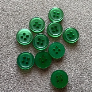 Könnte beinhalten: Zehn grüne Plastikknöpfe mit jeweils vier Löchern. Die Knöpfe sind in einem Halbkreis auf einer strukturierten grauen Oberfläche angeordnet.
