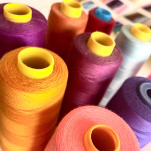 Può includere: Primo piano di una collezione di bobine di filo colorate. Le bobine sono disposte a semicerchio, con la bobina arancione al centro. Le bobine sono di diversi colori, tra cui viola, rosa, blu e arancione.