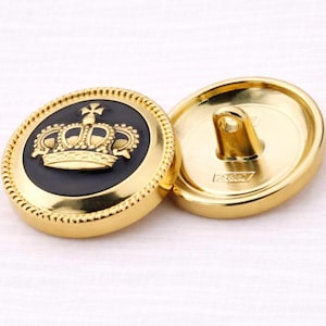 Peut inclure: Deux boutons dorés avec un motif de couronne. Les boutons ont un centre noir avec une couronne dorée et une bordure dorée. Un bouton est face visible et l'autre est face cachée, montrant la boucle pour la couture.