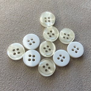 Puede incluir: Una colección de diez botones de plástico blanco, cada uno con cuatro agujeros. Los botones están dispuestos en forma de semicírculo.