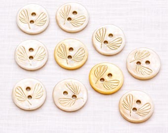 May include: A collection of ten round, off-white buttons with a leaf design. Each button has two holes and a carved leaf pattern in a lighter shade of gold. The buttons are arranged on a white, textured surface.