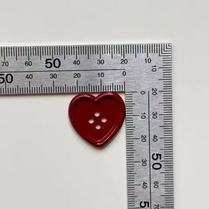 Pode incluir: Um bot&atilde;o vermelho em forma de cora&ccedil;&atilde;o com quatro orif&iacute;cios, com aproximadamente 2,5 cm de di&acirc;metro, est&aacute; posicionado perto de uma r&eacute;gua. O bot&atilde;o &eacute; vermelho brilhante e liso. A r&eacute;gua &eacute; prateada com marca&ccedil;&otilde;es pretas.