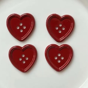 Pode incluir: Quatro bot&otilde;es em forma de cora&ccedil;&atilde;o, em vermelho profundo e brilhante. Cada bot&atilde;o tem quatro pequenos orif&iacute;cios para costurar. Os bot&otilde;es est&atilde;o dispostos numa superf&iacute;cie branca, perfeitos para projetos de artesanato ou para adicionar um toque de amor.