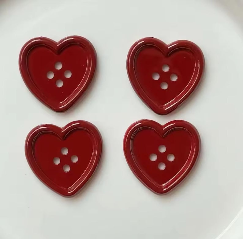 Pode incluir: Quatro bot&otilde;es em forma de cora&ccedil;&atilde;o, em vermelho profundo e brilhante. Cada bot&atilde;o tem quatro pequenos orif&iacute;cios para costurar. Os bot&otilde;es est&atilde;o dispostos numa superf&iacute;cie branca, perfeitos para projetos de artesanato ou para adicionar um toque de amor.