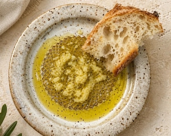 Pode incluir: Um prato de cerâmica salpicada contendo uma poça de azeite dourado misturado com alho picado e ervas, apresentando um padrão de grade texturizado na base. Uma fatia de pão de massa fermentada crocante e poroso repousa no azeite.