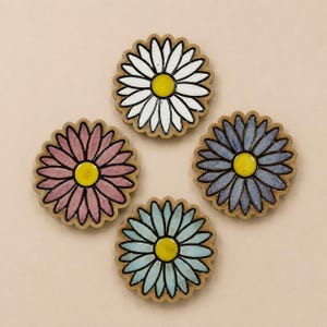 以下が含まれることがあります： デイジーの花の形をした4枚の円形セラミック製コースター。各コースターは鮮やかな黄色の中心部を持ち、その周りを白、ダスティピンク、ペリウィンクルブルー、ライトターコイズの長く細い花びらが囲んでいます。縁は自然な粘土色です。