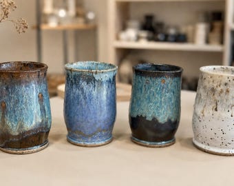 May include: Four ceramic tumblers arranged in a row. The first is brown and blue with a drip glaze. The second is a solid blue with a textured finish. The third is a dark blue and teal combination. The fourth is white with black speckles.