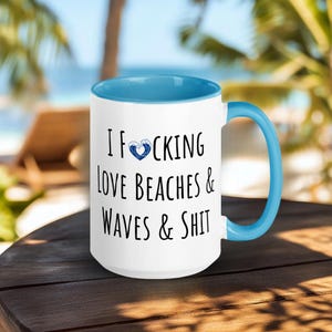 Puede incluir: Taza de café de cerámica blanca con borde y asa azules. La taza tiene un gráfico negro que dice "I F*CKING LOVE BEACHES & WAVES & SHIT" con un corazón de ola azul.