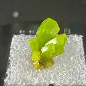 May include: A vibrant, lime-green mineral specimen with crystalline structures. The mineral is displayed in a clear, square case with a black frame, set against a neutral background. The crystals have a bright, almost fluorescent appearance.