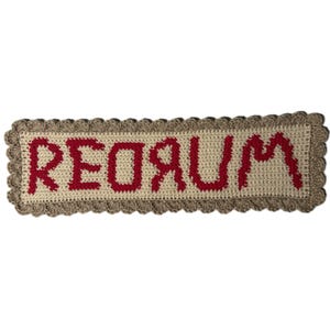 Puede incluir: Un letrero rectangular de ganchillo hecho a mano con la palabra "REDRUM" en letras rojas. El letrero tiene un fondo beige y un borde marrón festoneado. El texto está centrado en el letrero.