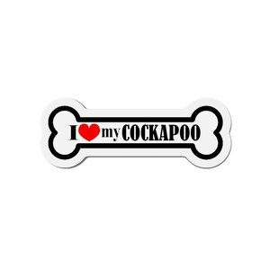 May include: A white bone-shaped sticker with black trim and the text "I ❤️ my COCKAPOO" in black. The heart is red. The sticker is designed to be placed on a car, laptop, or other surface.
