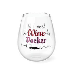 May include: A clear glass wine glass with a pink and red design that says "All I need is wine & my Docker". The glass has a black silhouette of a wine glass with a red wine spill and paw prints.