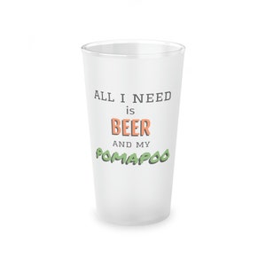Pode incluir: Copo de vidro branco fosco com o texto "All I need is beer and my Pom-a-poo" em preto, laranja e verde.