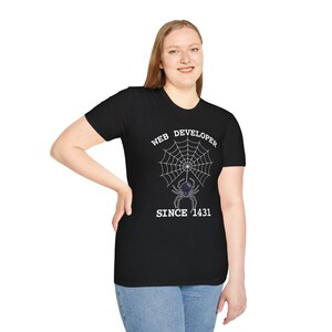 May include: Black t-shirt featuring a white spider web design and a purple spider. The text on the shirt reads "WEB DEVELOPER SINCE 1431". The t-shirt has a crew neck and is being worn by a person.