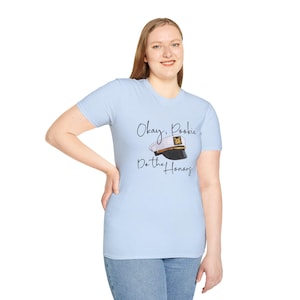 May include: Light blue t-shirt with the text "Okay, Pookie Do the Honors" and a graphic of a white and black captain's hat with a gold emblem. The shirt is made of a soft material.