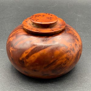 May include: A small, round, wooden container with a lid. The container and lid are a warm, reddish-brown color with a marbled grain pattern. The lid is flat with a small, circular handle.