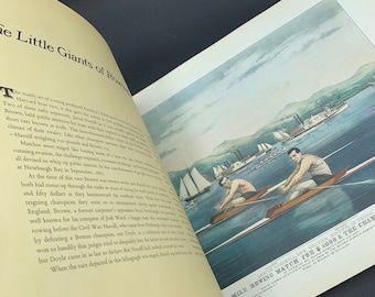 Op de afbeelding: Open boek met een vintage illustratie van een roeiwedstrijd. Twee mannen in een boot roeien op het water, met zeilboten en stoomboten op de achtergrond. De tekst op de pagina luidt "The Little Giants of Rowing."