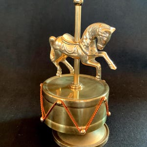 May include: A polished brass carousel horse figurine mounted on a central pole atop a cylindrical drum-shaped base. The base is decorated with thin orange cord detailing arranged in a zigzag pattern around the perimeter of the metal drum.