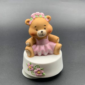 May include: A ceramic music box featuring a brown teddy bear wearing a pink tutu and a floral headband. The bear sits atop a white base decorated with pink flowers. The music box is a charming decorative item.