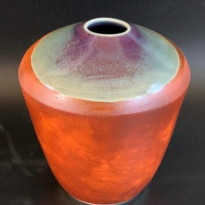 May include: A handmade ceramic vase with a wide, rounded body and a narrow opening. The vase features a gradient of colors, with a reddish-orange base, a green band, and a purple top. The interior is dark.