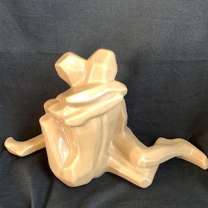 May include: A glossy, light beige ceramic sculpture depicting two abstract figures in a seated embrace. The piece features smooth, faceted surfaces and geometric forms, with one figure wrapping arms around the other in a unified, singular composition.