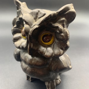 May include: A ceramic owl figurine with brown glaze and yellow eyes. The owl is perched on its feet with its wings folded.
