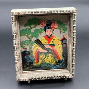 May include: Vintage framed artwork featuring a woman in a yellow kimono with red accents, holding a fan. The artwork is set against a backdrop of a garden scene with a tree and a pagoda. The frame is distressed white with a decorative edge.
