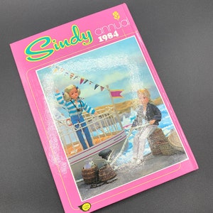 May include: A pink book with the title "Cindy annual 1984" on the cover. The cover features two dolls on a boat with a basket of fish and a rope. The boat is labeled "Adventure".