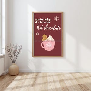 Könnte beinhalten: Gerahmter Druck mit burgunderfarbenem Hintergrund. Der Text „santa baby, it's time for hot chocolate“ ist zu sehen. Eine rosa Tasse mit Schneeflocken-Design enthält Schlagsahne und einen Lebkuchenmann.