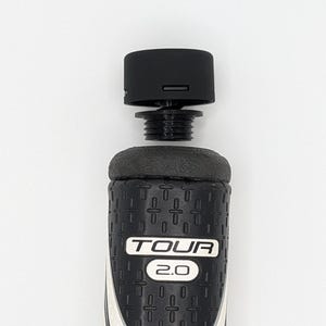 May include: A black and white golf club grip with the text "TOUR 2.0". The grip has a textured pattern and a white accent. A black cap is attached to the top of the grip.