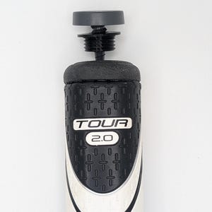 May include: A golf club grip with a black and white design. The grip features the text "TOUR 2.0" in white. The top of the grip has a black screw-on cap. The grip is designed for comfort and control during a golf swing.