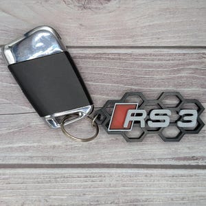 May include: Black car key next to a black and gray RS3 keychain. The keychain has a honeycomb design and the letters "RS3" in gray with a red background. The key has a silver top.