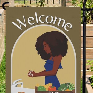 May include: A brown garden flag with the text "Welcome to my Happy Place!" in white. A cartoon illustration of a woman with brown skin and curly black hair washing her hands at a sink with a variety of vegetables in a wooden crate behind her.