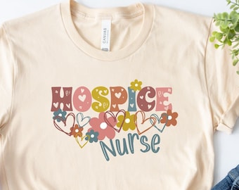 May include: A beige t-shirt with the text "Hospice Nurse" in colorful letters surrounded by flowers and hearts. The shirt is laid flat on a white surface with a pair of white sneakers, a wooden bead necklace, a brown tassel, and a pair of blue denim shorts with sunglasses and a lollipop.